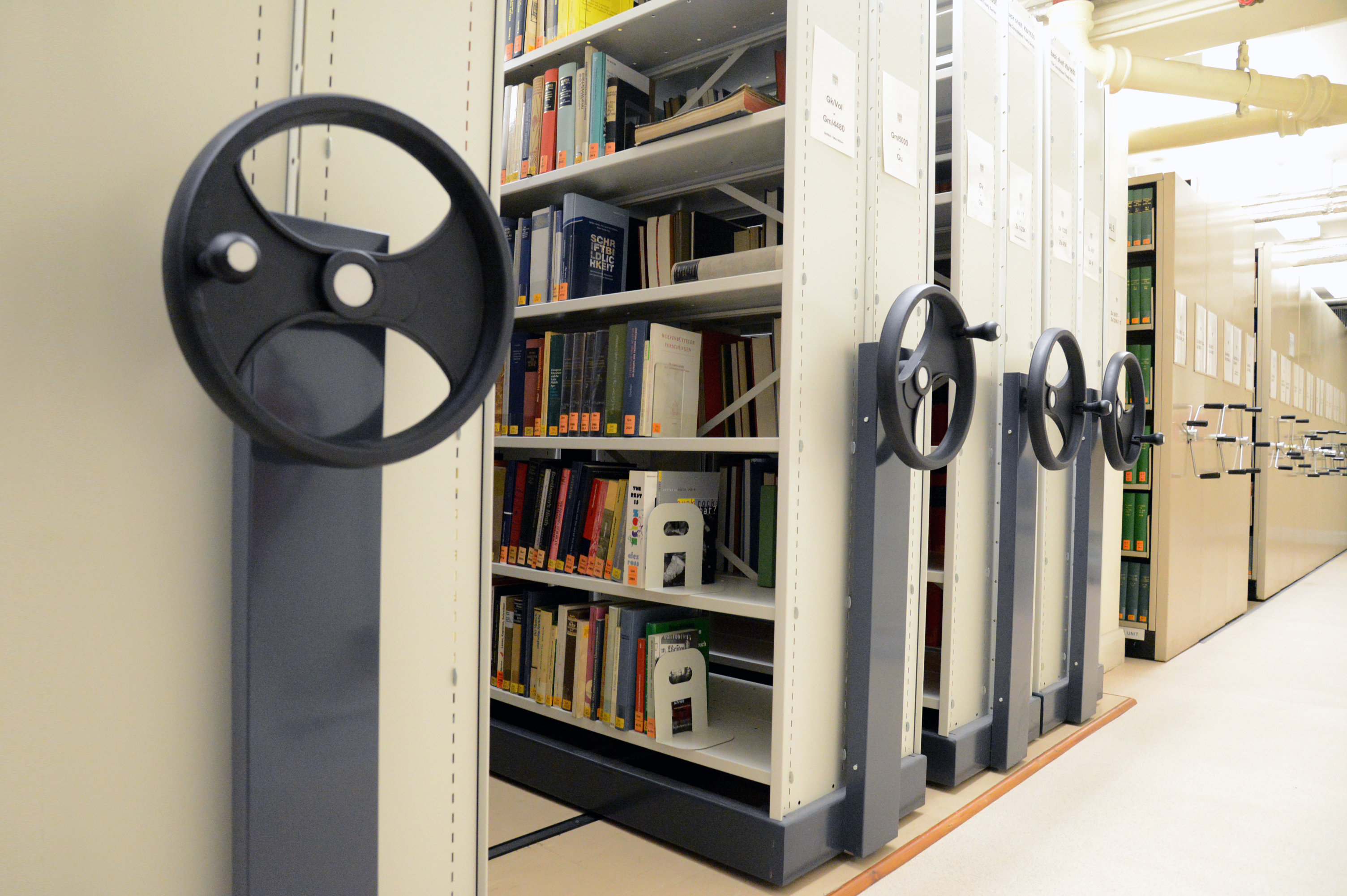 Movable library shelving units with books, the wheels to move the roller racking can be seen in the foreground