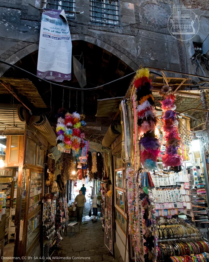 The Khan al-Harir (silk khan) in Damascus. Photo by Dosseman, 2008. The image shows a narrow street, partially built over, with stalls selling ribbons, pom-poms, and other goods on either side.