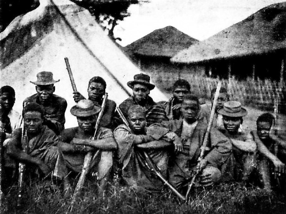 A group of African elephant hunters holding firearms, sitting in front of some tents and houses