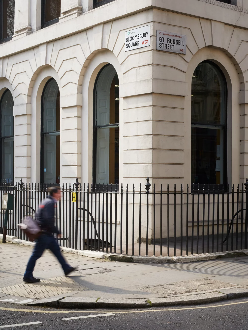 The corner of the GHIL building facing on Bloomsbury Square and Great Russell Street. A blurry man with a shoulder bag walks by in the direction of the British Museum.