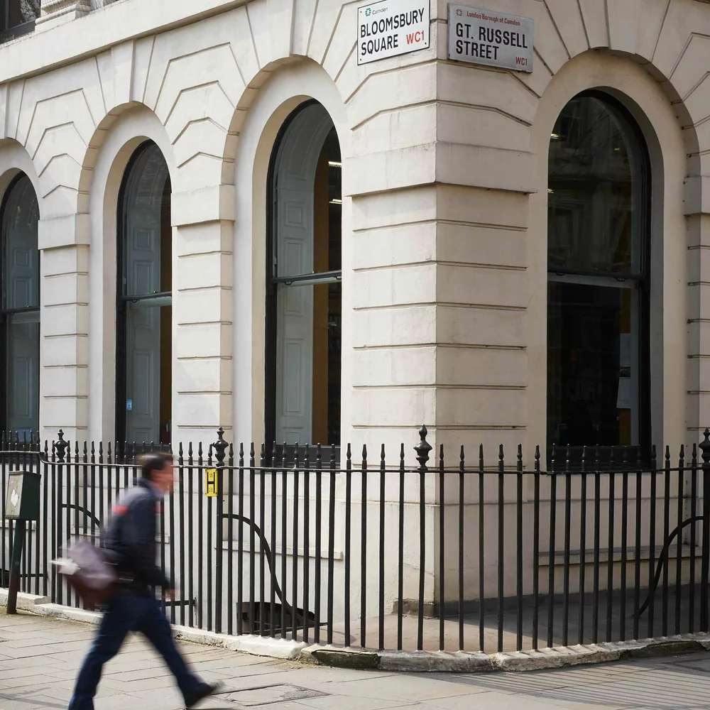 The corner of the GHIL building facing on Bloomsbury Square and Great Russell Street. A blurry man with a shoulder bag walks by in the direction of the British Museum.