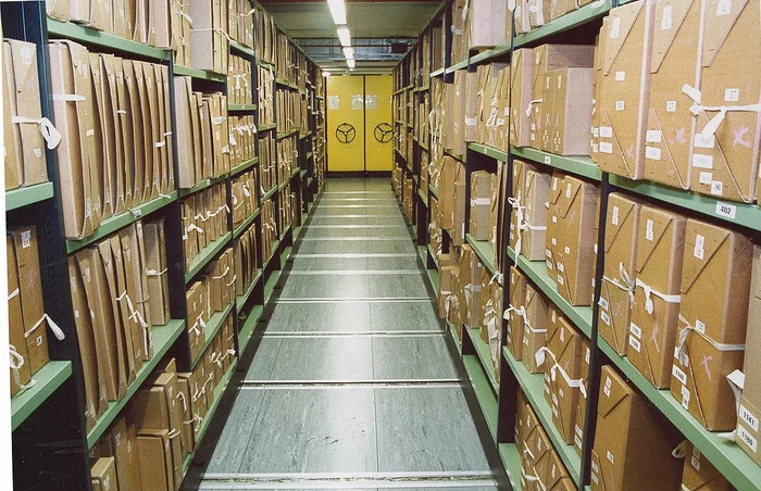 Documents stacks in a repository at The National Archives: archive boxes on green metal shelves, perspective narrowing to a bright yellow door at the end of the rows of shelves.