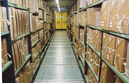 Documents stacks in a repository at The National Archives: archive boxes on green metal shelves, perspective narrowing to a bright yellow door at the end of the rows of shelves.
