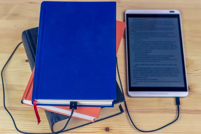A pile of three books next to an eBook reader, which is connected to one of the books by a USB cable.
