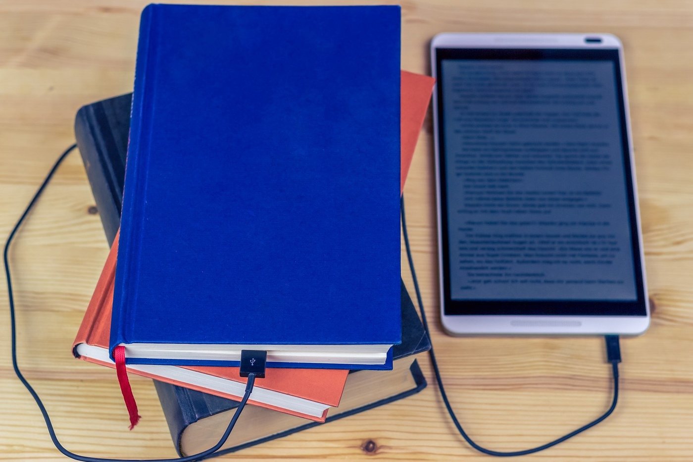 A pile of three books next to an eBook reader, which is connected to one of the books by a USB cable