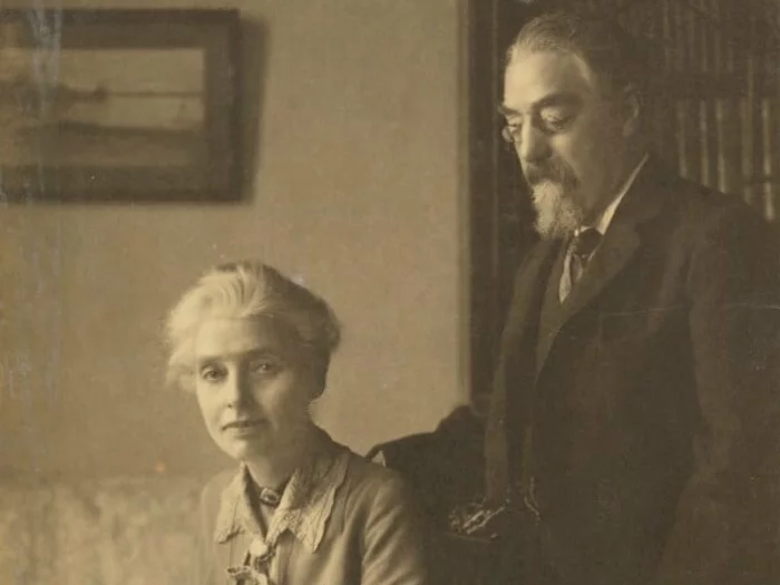 Black and white photograph of Beatrice Webb and Sidney James Webb, Baron Passfield. She is on the left of the image, seated, angled to the left, with a sheaf of papers in her hands, looking directly at the viewer. He is on the right of the image, standing behind her chair with his right hand on the back of the chair, looking down at her.