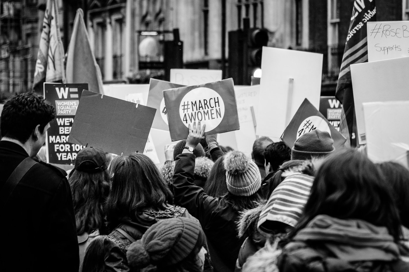 Black and white image of of a group of protesters, seen from behind. All are holding up placards, the most visible one in the cenbtre of the image reads 'March 4 Women'