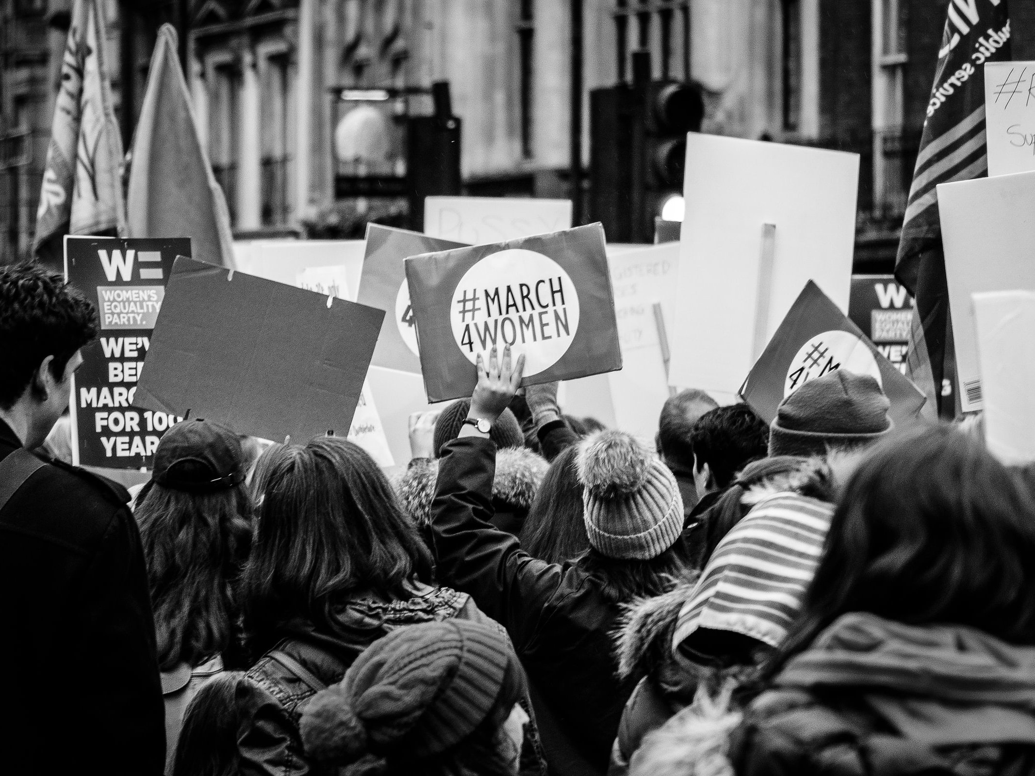 Black and white image of of a group of protesters, seen from behind. All are holding up placards, the most visible one in the cenbtre of the image reads 'March 4 Women'