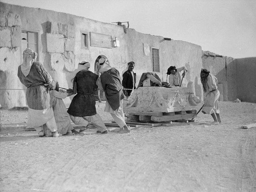 A group of seven workmen at Palmyra moving a massive capital on a wooden sledge by pulling it over wooden poles by means of a rope wrapped around the capital.