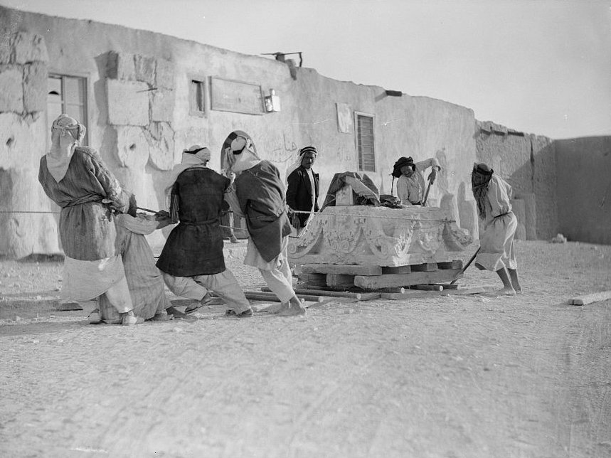 A group of seven workmen at Palmyra moving a massive capital on a wooden sledge by pulling it over wooden poles by means of a rope wrapped around the capital.