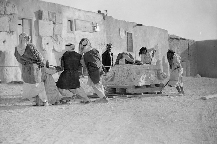 A group of seven workmen at Palmyra moving a massive capital on a wooden sledge by pulling it over wooden poles by means of a rope wrapped around the capital.