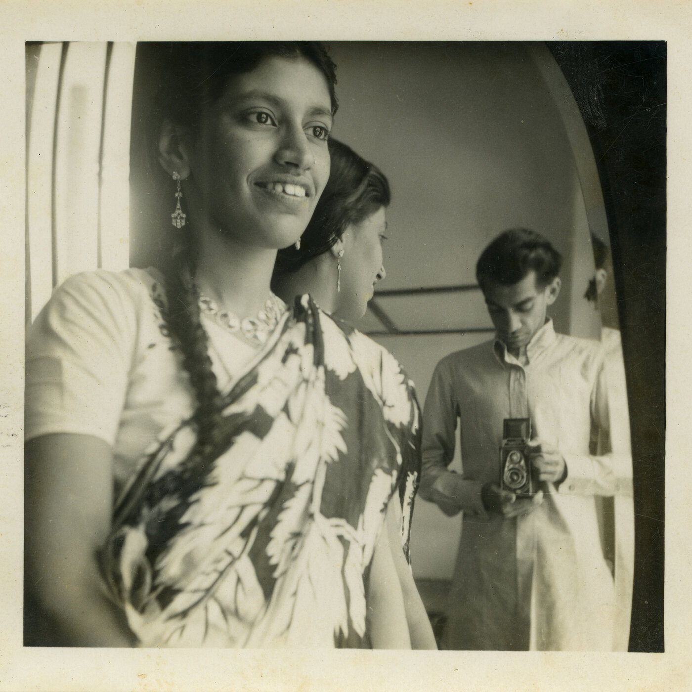 Haleema Hashim and Hashim Usman, Self-portrait, c. 1955. Black and white photograph. The woman is standing in front of a mirror, so can be seen from the front and the side. The man, who is the photographer, can only seen in the mirror. He is looking down at the camera. She is looking at him, so neither gaze is bent upon the viewer.