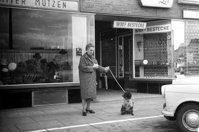 A black and white image of an older lady with her spaniel, in front of a rank of shops on Hamburger Strasse in Hamburg, 1965, featuring cutlery and men's hats.