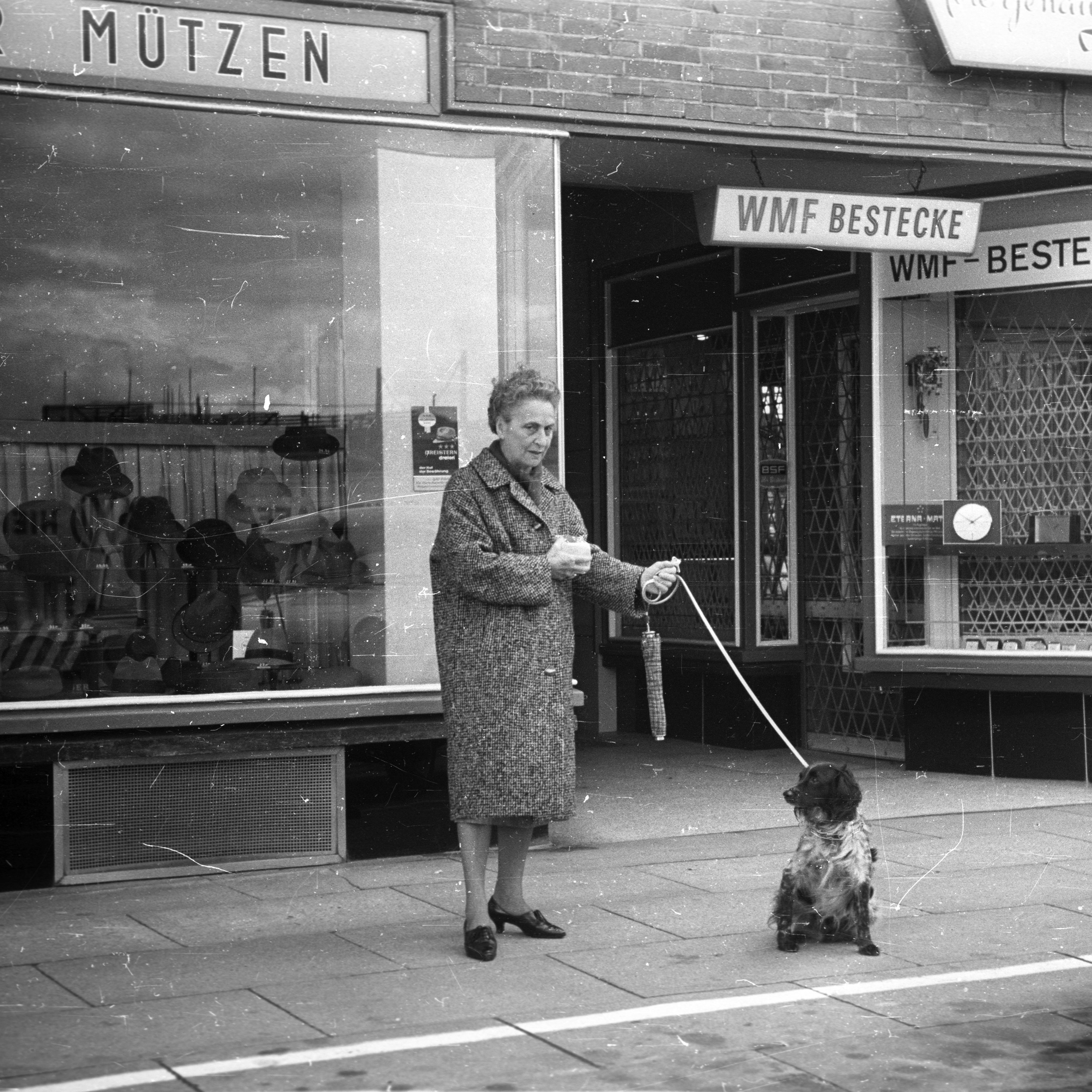 A black and white image of an older lady with her spaniel, in front of a rank of shops on Hamburger Strasse in Hamburg, 1965, featuring cutlery and men's hats