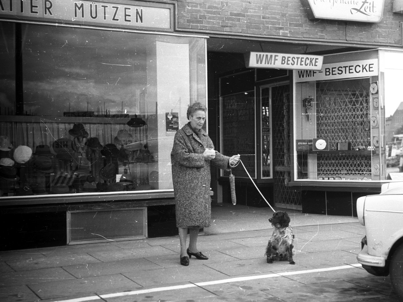 A black and white image of an older lady with her spaniel, in front of a rank of shops on Hamburger Strasse in Hamburg, 1965, featuring cutlery and men's hats
