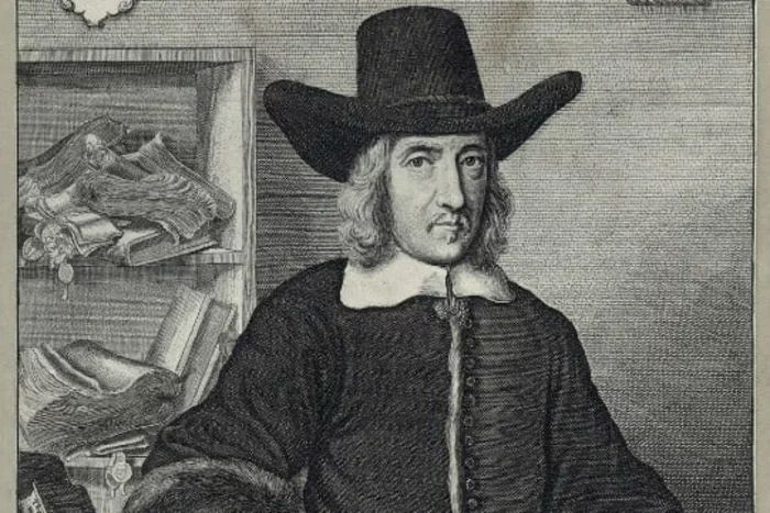 Etching of Sir William Dugdale, by Wenceslaus Hollar, 1656. The image shows Dugdale seated, with a desk covered with books and scrolls to his right. His hand rests lightly on a scroll, which sits next to an inkwell. He is wearing a coat with large fur cuffs. His white shirt collar and the ruffs at the ends of his sleeves are visible. He has on his head a large hat. Behind him on his right side are shelves with more scrolls.