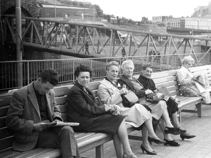 A black and white photograph of St. Pauli-Landungsbrücken, Hamburg, 1965, featuring several older ladies and one younger man, all sitting on benches.