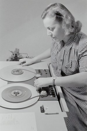 Black and white image of a woman in a checkered dress using an Austrovox tape recorder, 1947