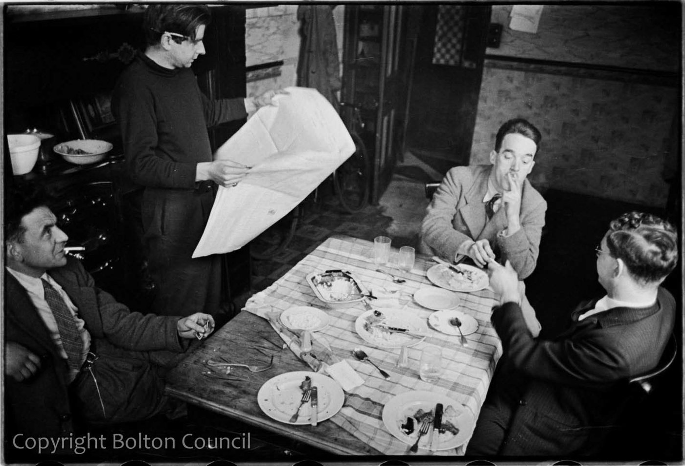 A black and white photograph of a group of four men from the Mass Observation Project, sitting and smoking around a small dining table, on which the rests of a meal can be seen.
