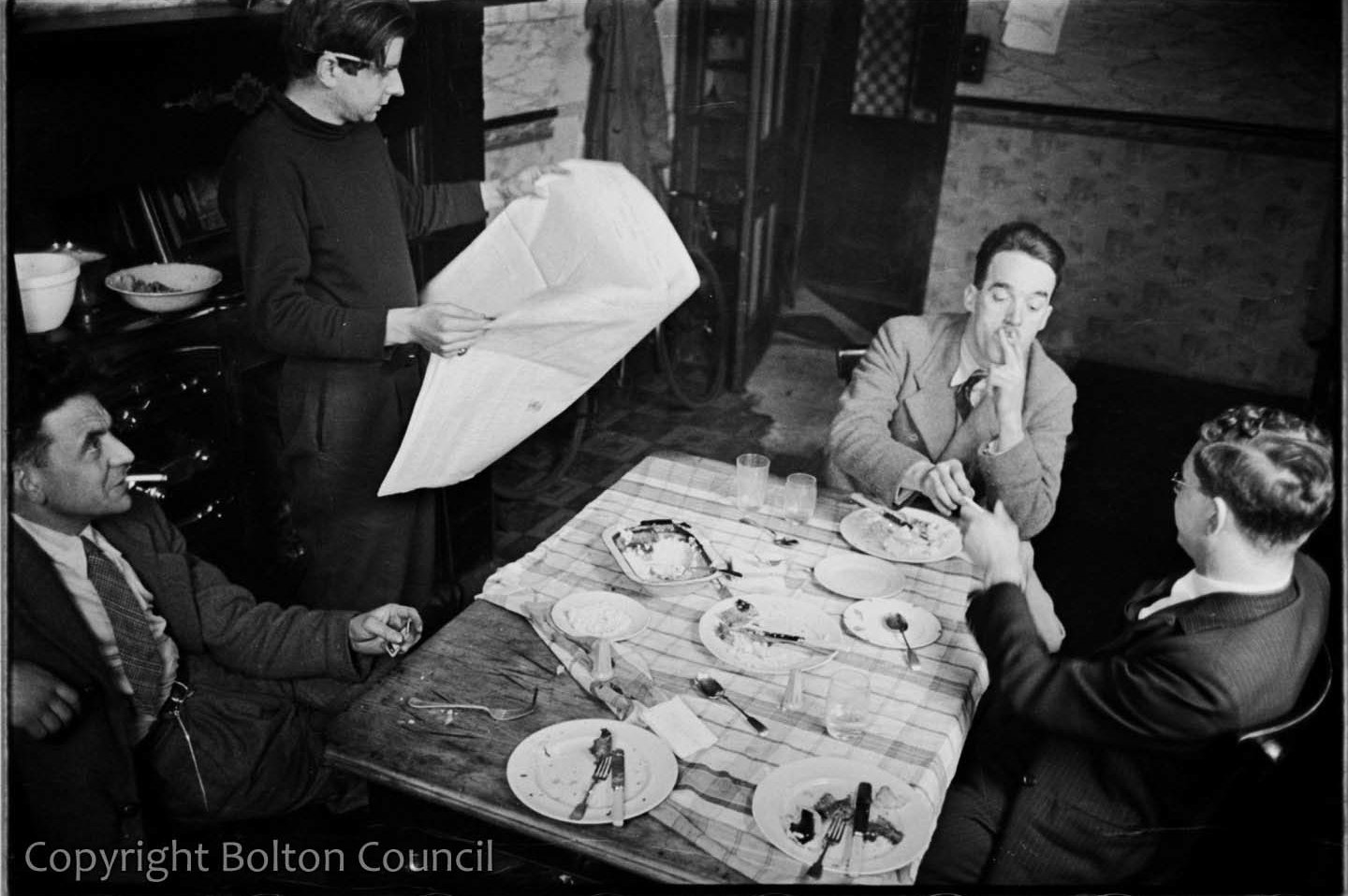 A black and white photograph of a group of four men from the Mass Observation Project, sitting and smoking around a small dining table, on which the rests of a meal can be seen.