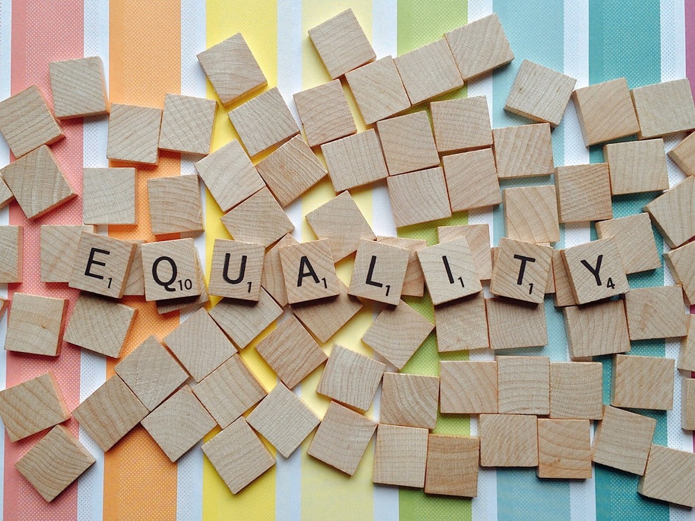 Scrabble tiles spelling out the word 'equality', on a pile on blank tiles on top of a rainbow stripe background