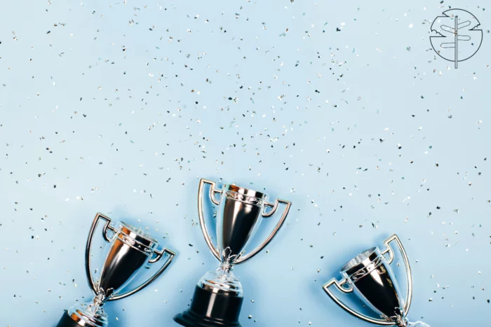 Three small silver trophies on black bases, on a blue background, with silver confetti falling down on them.