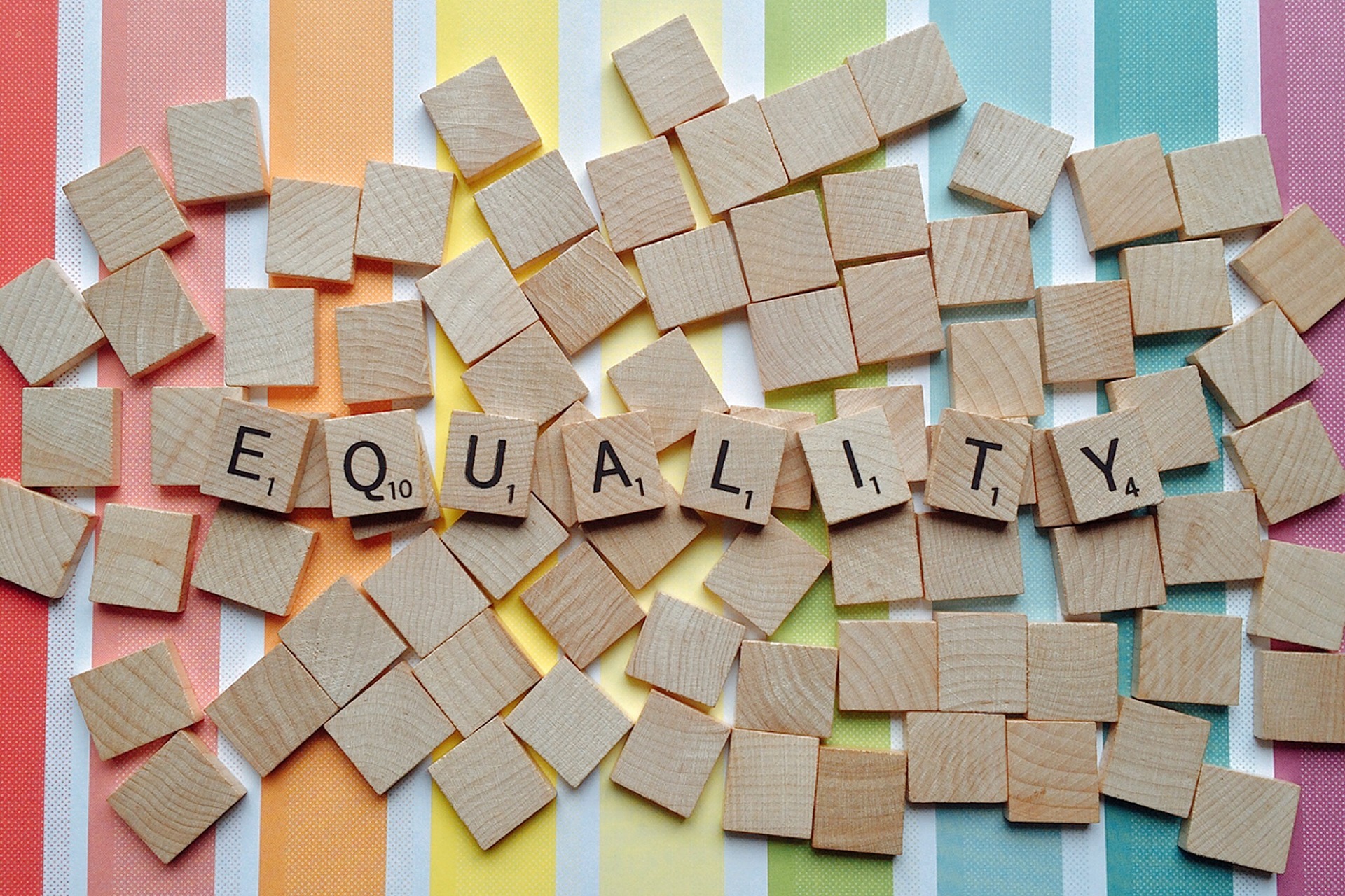 Scrabble tiles spelling out the word 'equality', on a pile on blank tiles on top of a rainbow stripe background