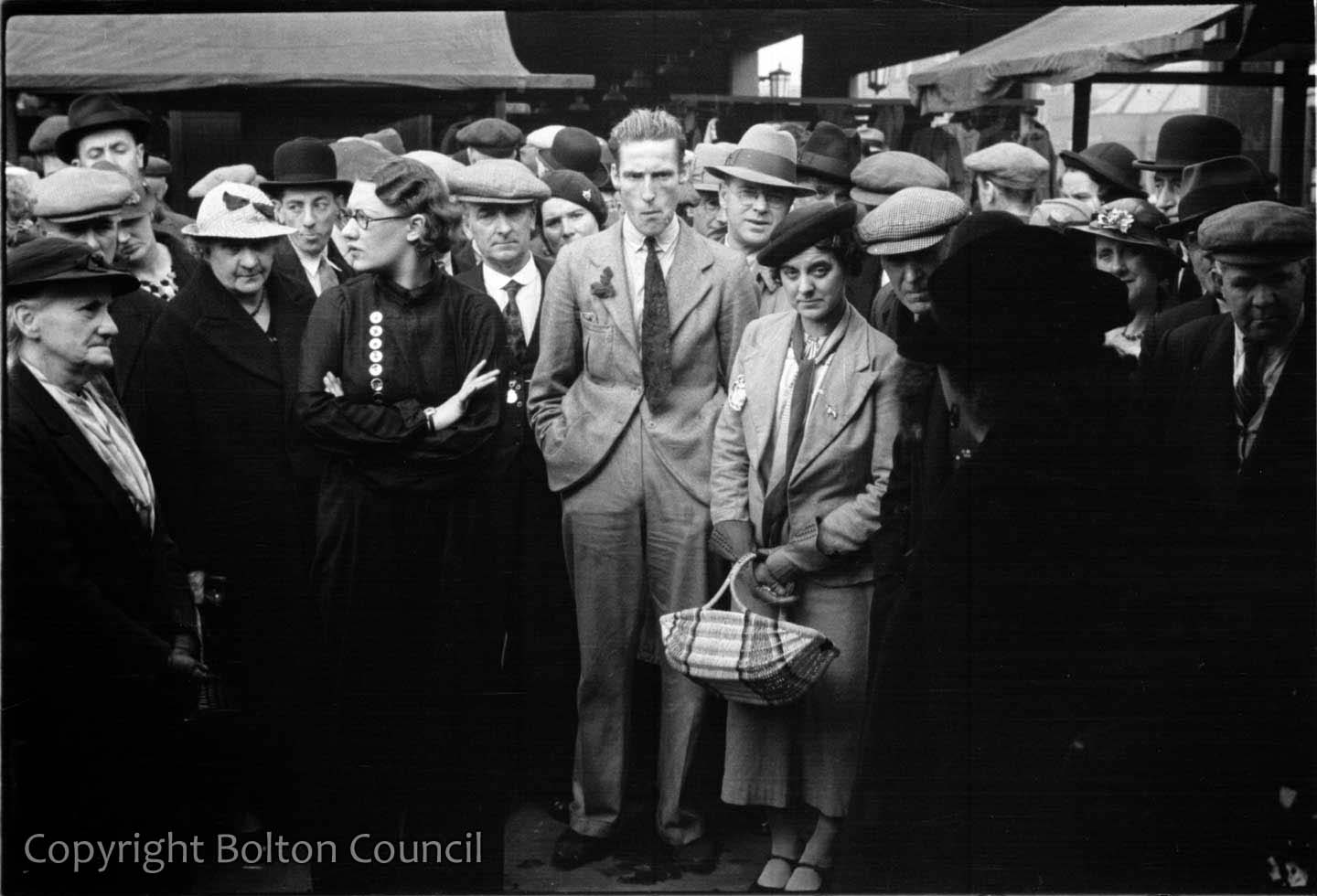 A black and white photograph of shoppers surrounding a stall in Bolton’s open market. A woman in the foreground holds a shopping basket.