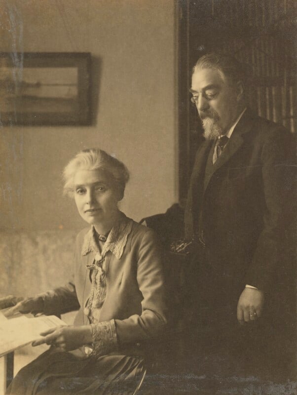 Black and white photograph of Beatrice Webb and Sidney James Webb, Baron Passfield. She is on the left of the image, seated, angled to the left, with a sheaf of papers in her hands, looking directly at the viewer. He is on the right of the image, standing behind her chair with his right hand on the back of the chair, looking down at her.