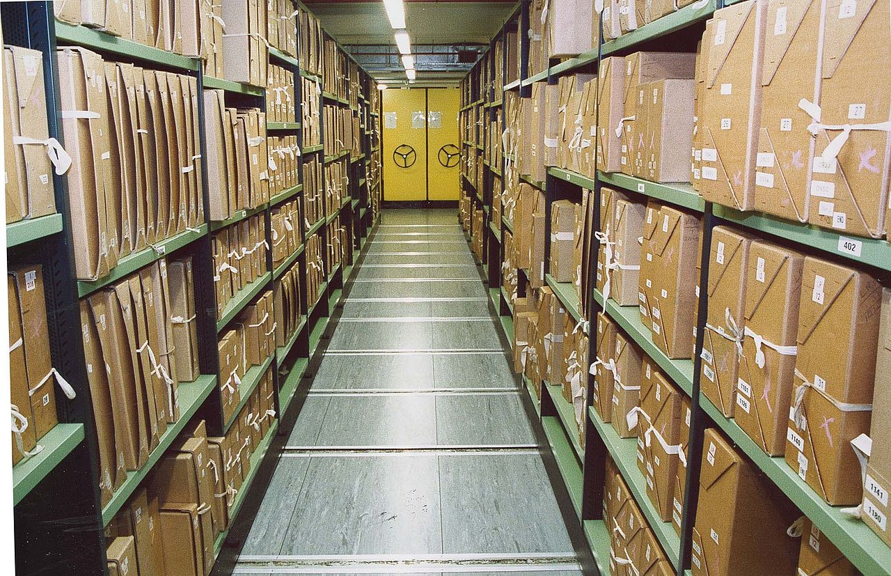 Documents stacks in a repository at The National Archives: archive boxes on green metal shelves, perspective narrowing to a bright yellow door at the end of the rows of shelves.