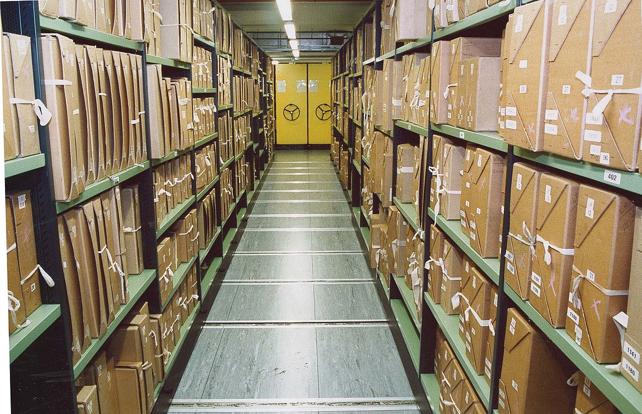 Documents stacks in a repository at The National Archives: archive boxes on green metal shelves, perspective narrowing to a bright yellow door at the end of the rows of shelves